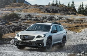 Weißer Subaru Outback fährt auf felsigen Gelände mit Tannen und schneebedeckten Bergen im Hintergrund