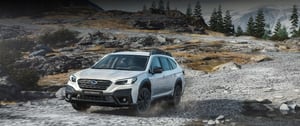 Weißer Subaru Outback fährt auf felsigen Gelände mit Tannen und schneebedeckten Bergen im Hintergrund
