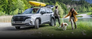 Silberner Subaru Forester mit gelbem Kajak auf dem Dach mit Bergen im Hintergrund und zwei Personen mit Hund
