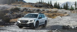 Weißer Subaru Outback fährt auf felsigen Gelände mit Tannen und schneebedeckten Bergen im Hintergrund