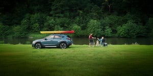 Subaru Crosstrek mit Kajak auf dem Dach links neben zwei Personen auf Klappstühlen sitzend. Waldiger Hintergrund.